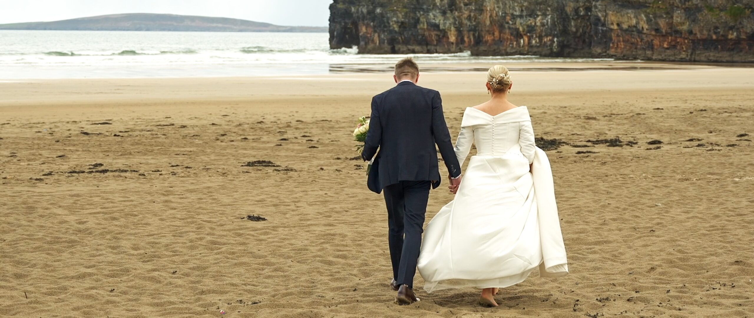 Newlywed couple holding hands and kissing while walking on Ballybunion beach after their wedding ceremony