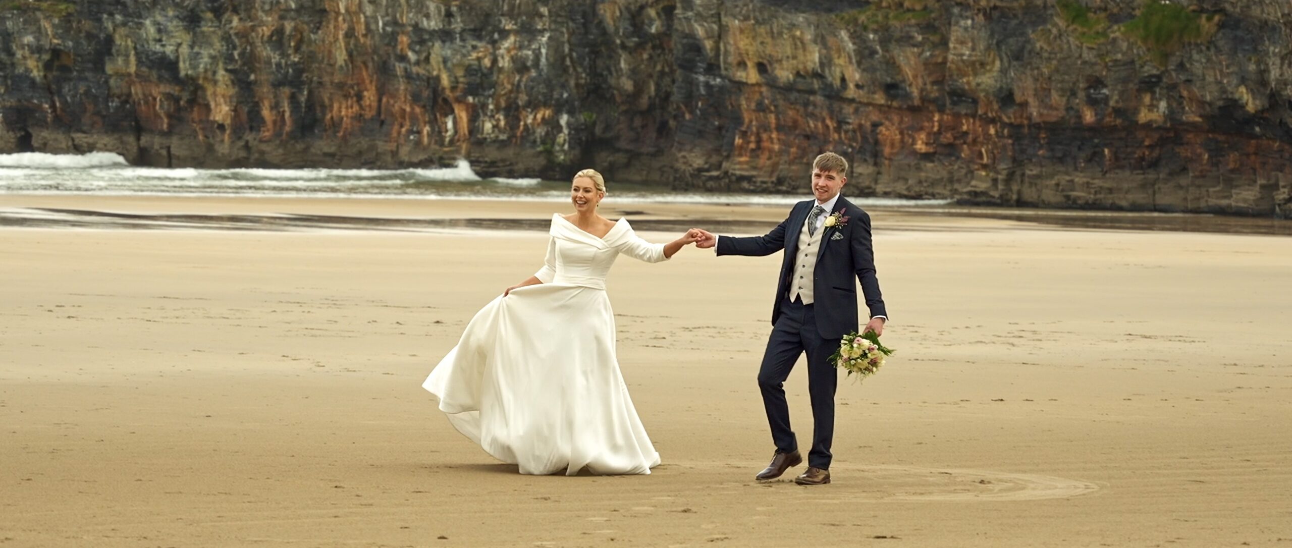 Newlywed couple holding hands and kissing while walking on Ballybunion beach after their wedding ceremony