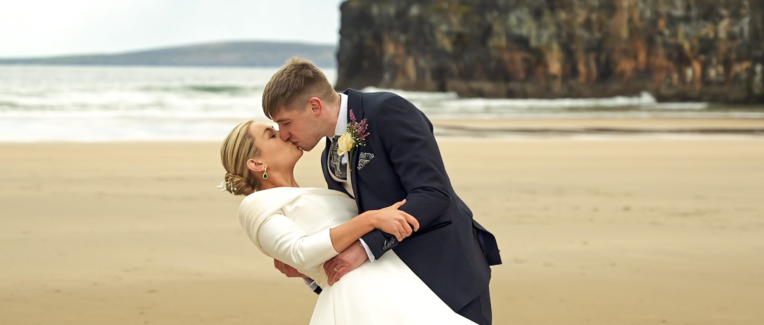 Newlywed couple holding hands and kissing while walking on Ballybunion beach after their wedding ceremony